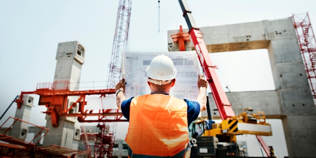 Construction worker holding up construction drawings on site with crane in background.